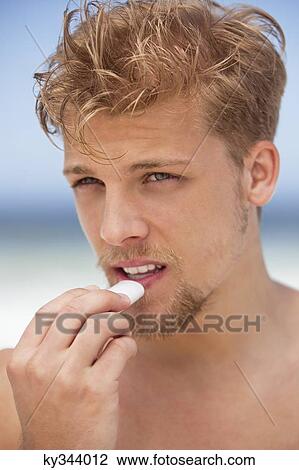 Stock Image - Young man applying moisturizer on his lips. Fotosearch