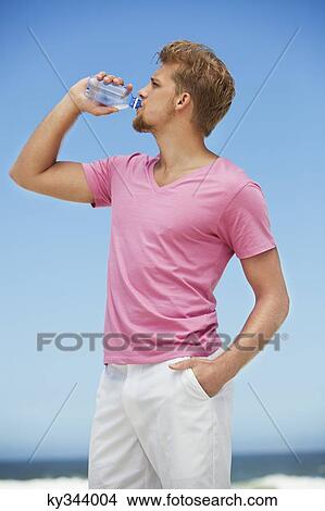 Picture - Young man drinking water from a water bottle on the beach. Fotosearch