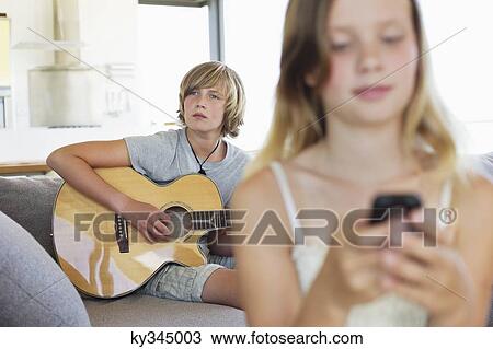 Stock Image - Teenage boy playing a guitar while his sister using a mobile phone. Fotosearch