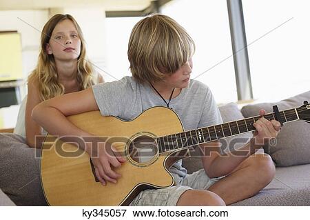 Teenage boy playing a guitar with her sister standing behind him listening View Large Photo Image Stock Photo - Teenage boy playing a guitar with her sister standing behind him listening. Fotosearch