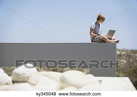 Stock Photo - Teenage boy sitting at the edge of a terrace and using a laptop. Fotosearch