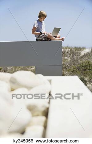 Teenage boy sitting at the edge of a terrace and using a laptop View Large Photo Image Stock Photograph - Teenage boy sitting at the edge of a terrace and using a laptop. Fotosearch