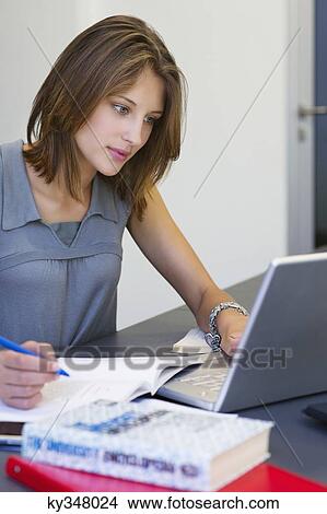 Picture - Beautiful woman writing in book while using laptop in classroom. Fotosearch