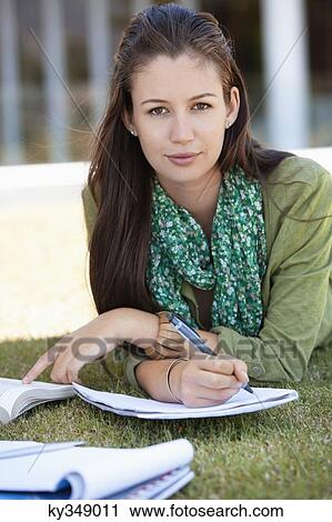 Stock Image - Portrait of young woman lying on grass and studying in a campus. Fotosearch