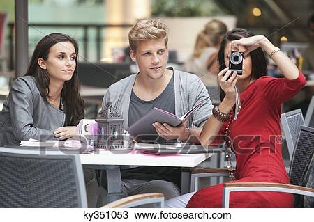 Three young friends sitting in a restaurant with one woman using camera View Large Photo Image Stock Image - Three young friends sitting in a restaurant with one woman using camera. Fotosearch
