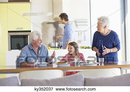 Girl sitting with her parents at a dining table and her father cooking food in the background View Large Photo Image Stock Photo - Girl sitting with her parents at a dining table and her father cooking food in the background. Fotosearch