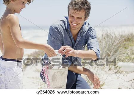 Stock Photograph - Mid adult man with his son cleaning the beach. Fotosearch