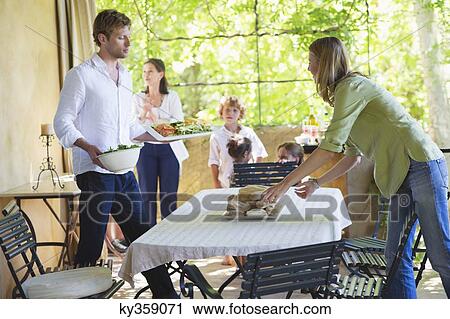 Couple arranging food on dining table with family in the background View Large Photo Image Stock Image - Couple arranging food on dining table with family in the background. Fotosearch