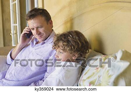 Picture - Father talking over mobile phone with little boy sitting with him on couch. Fotosearch