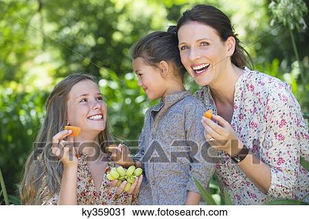Stock Image - Happy mother eating fruits with her two daughters outdoors. Fotosearch