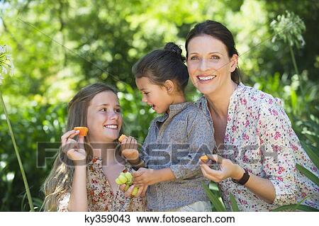 Stock Image - Happy mother eating fruits with her two daughters outdoors. Fotosearch