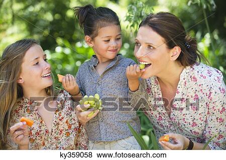 Happy mother eating fruits with her two daughters outdoors View Large Photo Image Stock Photo - Happy mother eating fruits with her two daughters outdoors. Fotosearch