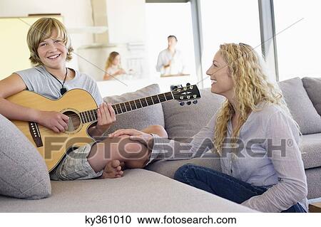 Teenage boy playing a guitar with his mother sitting near him and smiling View Large Photo Image Stock Image - Teenage boy playing a guitar with his mother sitting near him and smiling. Fotosearch
