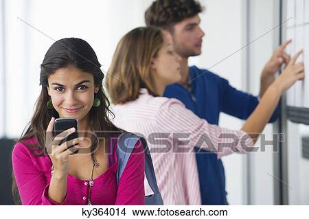 Picture - Portrait of university students using a mobile phone while friends checking for test results in bulletin board. Fotosearch