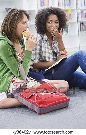Stock Photo - University student looking at her friend eating food in a library. Fotosearch