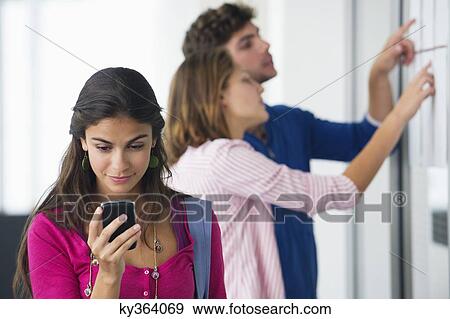 University students checking for test results in bulletin board while woman checking text message on mobile phone View Large Photo Image Stock Photo - University students checking for test results in bulletin board while woman checking text message on mobile phone. Fotosearch