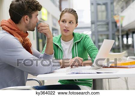 Stock Image - University students using laptop and looking at each other in lobby. Fotosearch