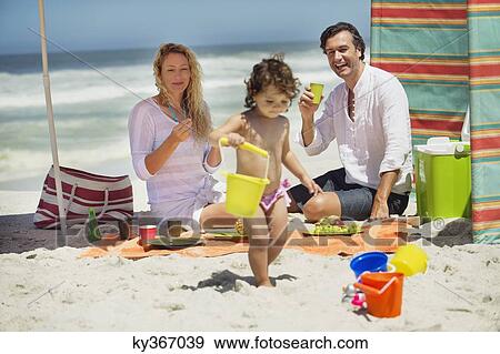 Stock Photo - Girl playing on the beach with her parents sitting behind her. Fotosearch
