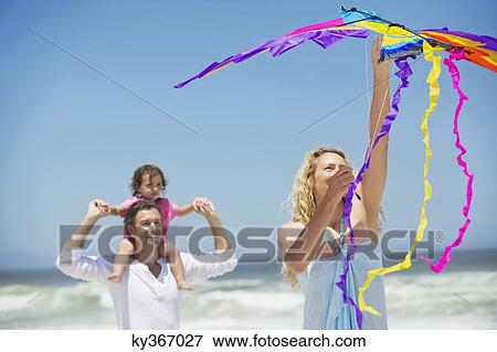 Little girl sitting on father's shoulder while mother flying shaped kite on the beach View Large Photo Image Stock Photo - Little girl sitting on father's shoulder while mother flying shaped kite on the beach. Fotosearch