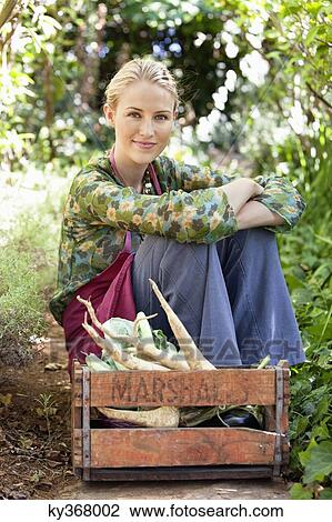 Stock Image - Smiling woman sitting near a crate of vegetables. Fotosearch