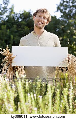 Stock Image - Young man in scarecrow dressing and holding a blank placard in a field. Fotosearch