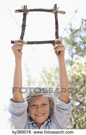 Stock Photograph - Portrait of a cute little boy holding frame of driftwood with arms raised outdoors. Fotosearch