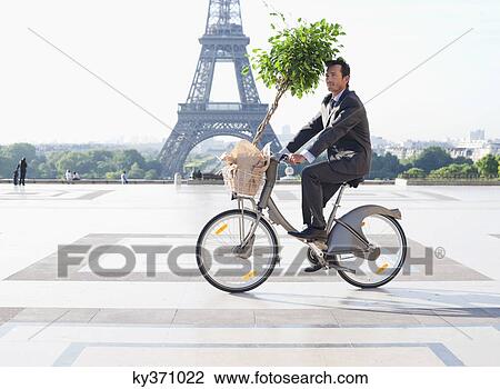 Stock Image - Businessman carrying a plant on a bicycle with the Eiffel Tower in the background, Paris, Ile-de-France, France. Fotosearch