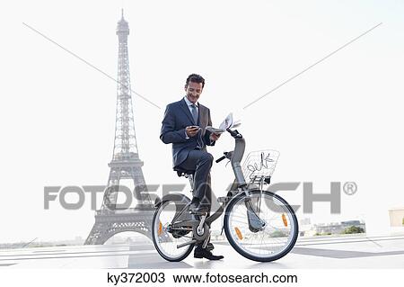 Stock Image - Businessman holding a newspaper and a mobile on a bicycle with the Eiffel Tower in the background, Paris, Ile-de-France, France. Fotosearch