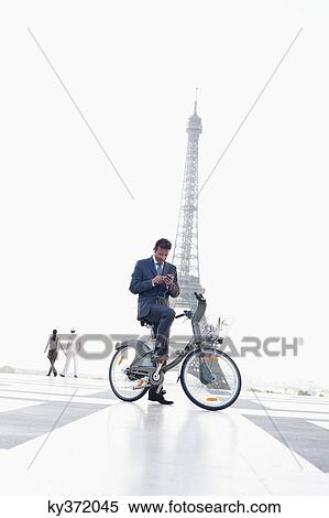 Stock Photography - Businessman operating a mobile phone on a bicycle with the Eiffel Tower in the background, Paris, Ile-de-France, France. Fotosearch