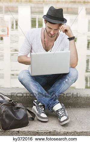 Stock Photograph - Man sitting on the ledge of a canal and using a laptop, Paris, Ile-de-France, France. Fotosearch