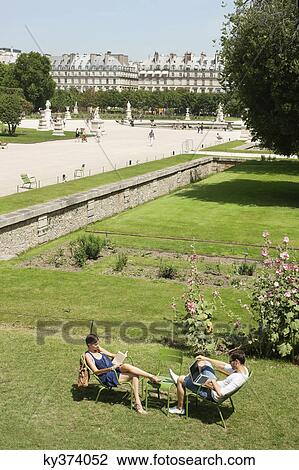 Stock Image - Man using a laptop with a woman reading a magazine in garden, Jardin des Tuileries, Paris, Ile-de-France, France. Fotosearch