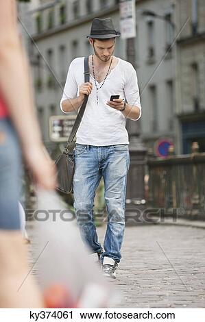 Stock Image - Man walking on the road and text messaging with a mobile phone, Paris, Ile-de-France, France. Fotosearch