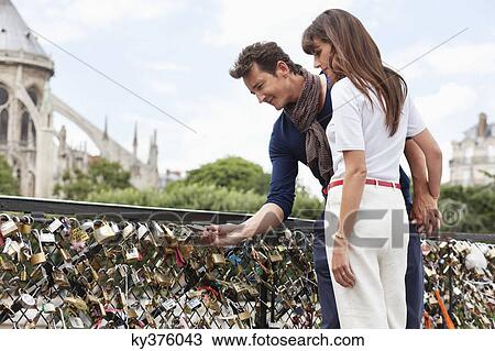 Stock Image - Couple locking a padlock of love on a bridge, Pont des Arts, Notre Dame de Paris, Paris, Ile-de-France, France. Fotosearch