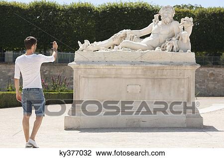 Stock Image - Man taking a picture of a sculpture in a garden, Jardin des Tuileries, Paris, Ile-de-France, France. Fotosearch