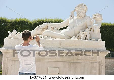 Stock Image - Man taking a picture of a sculpture in a garden, Jardin des Tuileries, Paris, Ile-de-France, France. Fotosearch