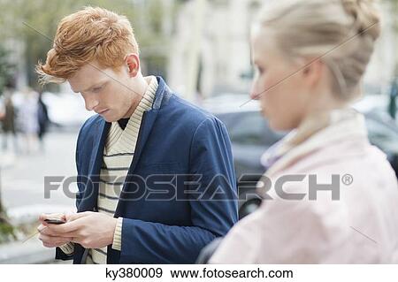 Stock Photo - Man text messaging on a mobile phone with her girlfriend beside him. Fotosearch
