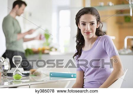 Woman smiling with her husband preparing food in the background View Large Photo Image Stock Photo - Woman smiling with her husband preparing food in the background. Fotosearch