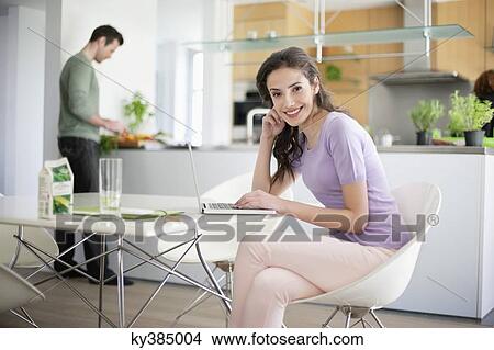 Woman using a laptop with her husband preparing food in the background View Large Photo Image Picture - Woman using a laptop with her husband preparing food in the background. Fotosearch