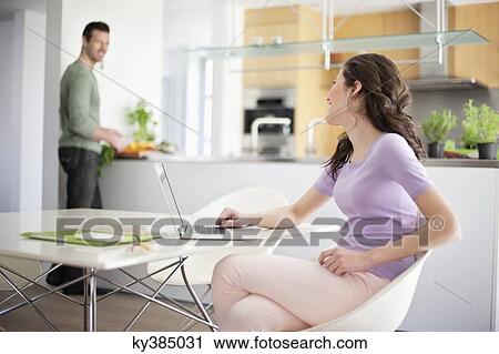Stock Image - Woman using a laptop with her husband preparing food in the background. Fotosearch