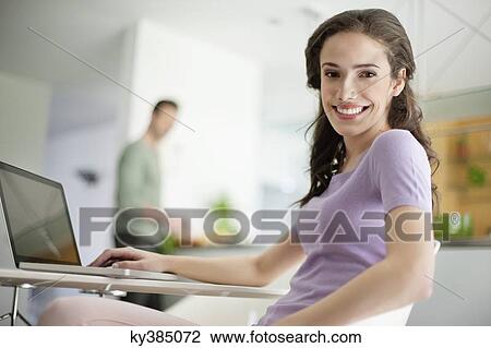 Woman using a laptop with her husband preparing food in the background View Large Photo Image Stock Image - Woman using a laptop with her husband preparing food in the background. Fotosearch