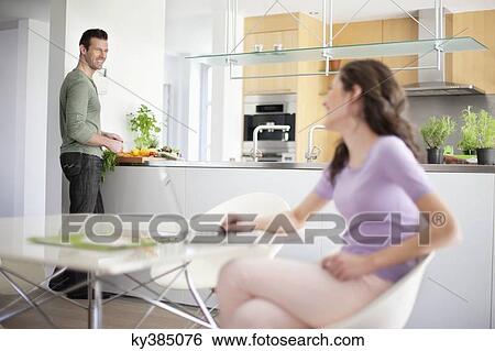 Woman using a laptop with her husband preparing food in the background View Large Photo Image Stock Photograph - Woman using a laptop with her husband preparing food in the background. Fotosearch