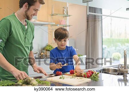 Picture - Man looking at his son cutting vegetables in the kitchen. Fotosearch