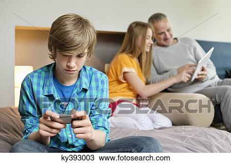 Teenage boy using mobile phone with his father and sister using digital tablet in background View Large Photo Image Stock Image - Teenage boy using mobile phone with his father and sister using digital tablet in background. Fotosearch