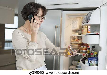 Stock Image - Elderly woman checking food items in a refrigerator and talking on a mobile phone. Fotosearch