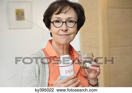Stock Image - Elderly woman holding bottle of food supplement in the kitchen. Fotosearch