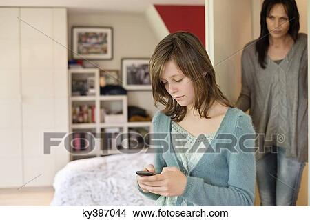 Girl text messaging on mobile phone with her mother watching her from behind the door View Large Photo Image Picture - Girl text messaging on mobile phone with her mother watching her from behind the door. Fotosearch