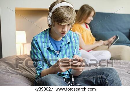 Stock Photo - Teenage boy listening to music on iPod with his sister using digital tablet. Fotosearch