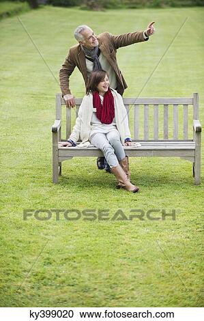 Stock Image - Man sitting with his daughter on a bench and pointing in a park. Fotosearch