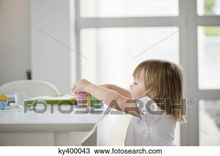 Stock Image - Cute little girl picking up toy tea set from a dining table. Fotosearch