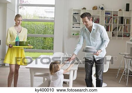 Stock Image - Woman serving food with her husband and daughter holding plates at dining table. Fotosearch